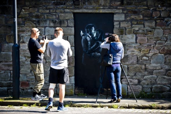 People photograph and film the latest officially confirmed Banksy artwork, named Mobile Lovers, featuring a man and a woman embraced and looking at their mobile phones, which is sprayed onto a black wooden board in a blocked off doorway on Clement Street, Bristol, within sight of the city's bustling centre. PRESS ASSOCIATION Photo. Picture date: Tuesday April 15, 2014. The latest artwork to be officially confirmed by Banksy has been tracked down to a street in the street artist's hometown. It comes days after a piece depicting three 1950s-style agents listening in on conversations in a telephone box appeared on a house in Cheltenham, Gloucestershire. Website streetartnews described the Bristol piece as 