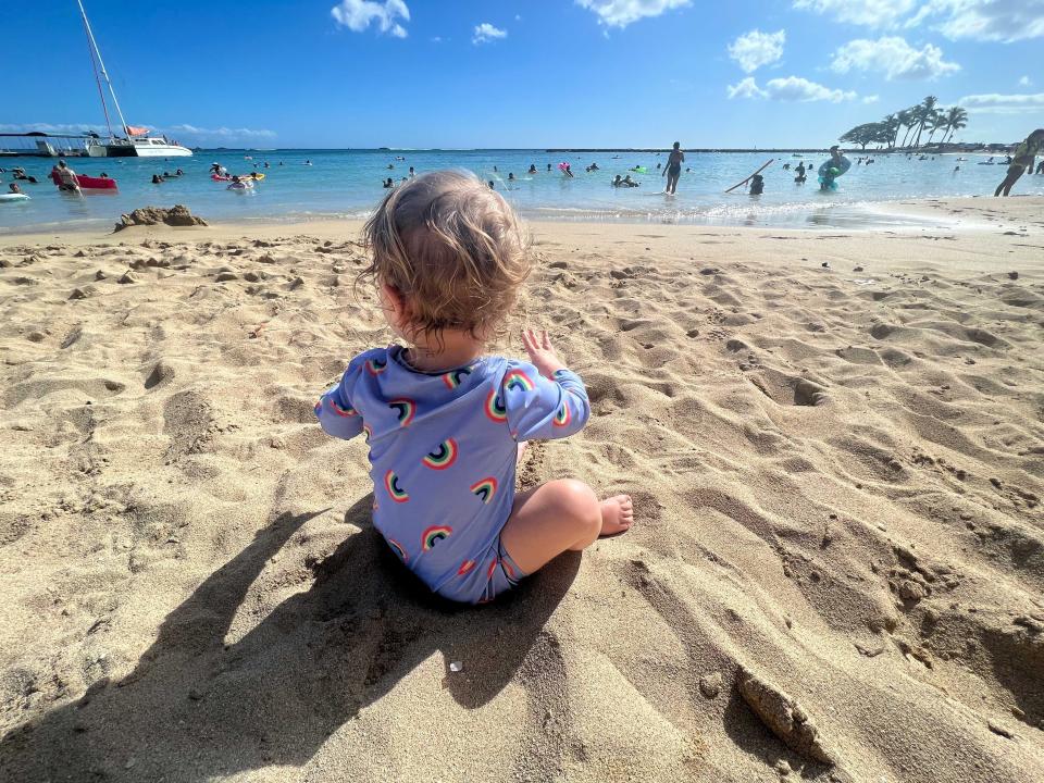 A baby sitting on the beach facing the ocean.