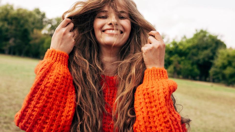 positive young woman playing with her long red hair wearing an orange knitted sweater posing on the nature background pretty female playing with her hair outdoors in the park