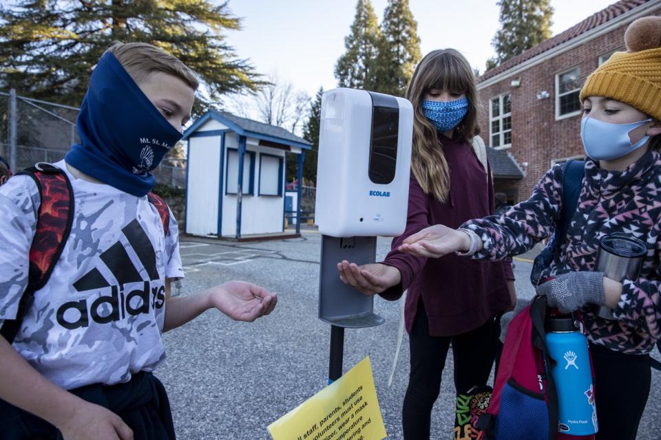 Students in masks hold their palms out to a hand sanitizer dispenser