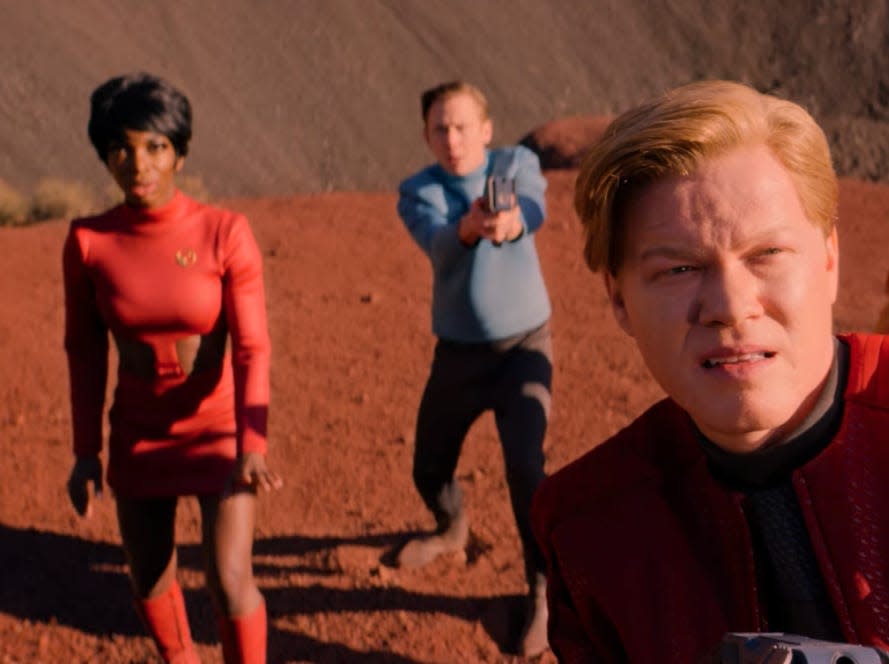 a group of people dressed in star trek-like red, blue, and yellow bright colored shirts, standing on the sand and cowering away from something