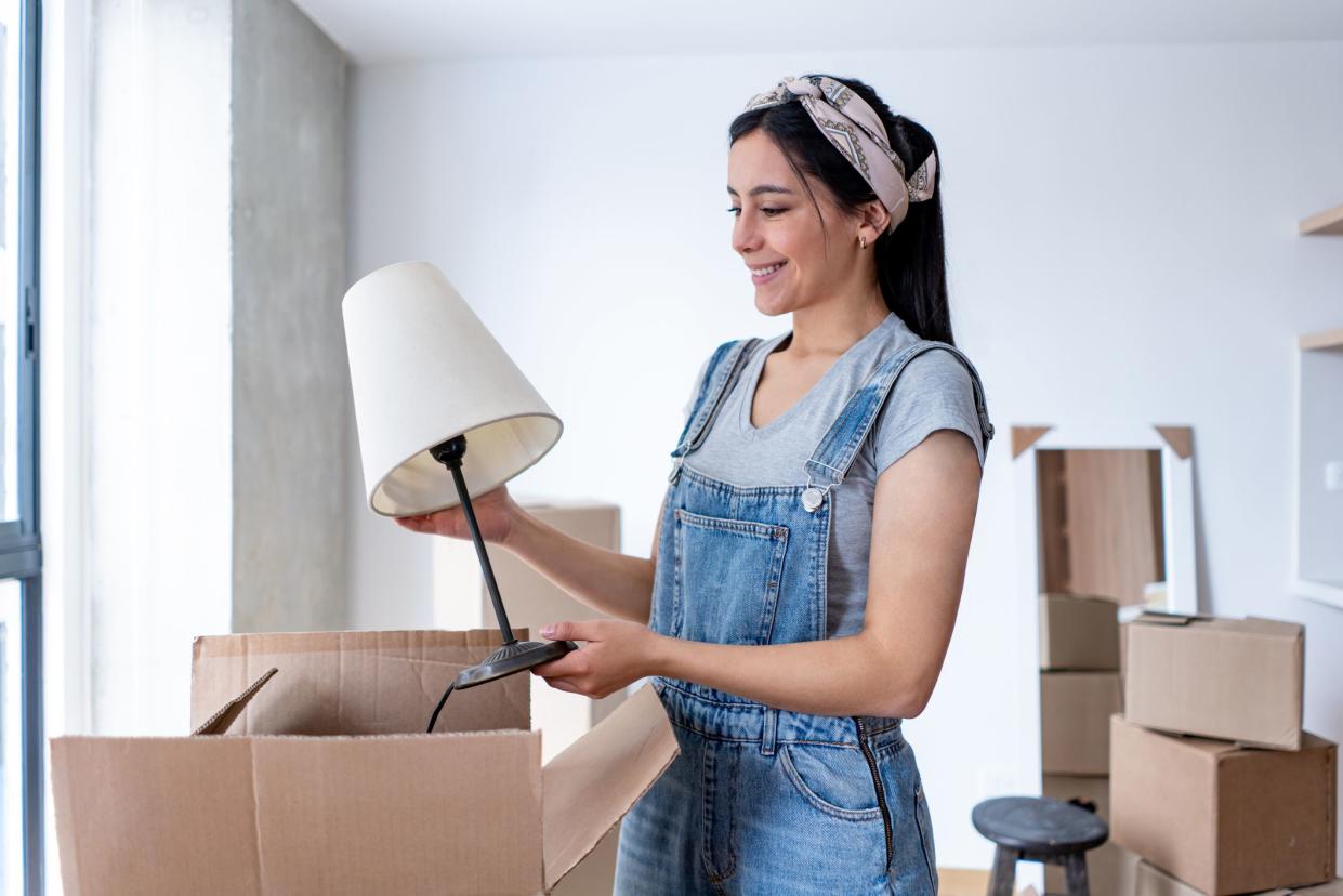 Young beautiful cheerful woman packing a lamp in a box ready to move to a new place looking very happy - New beginnings concepts
