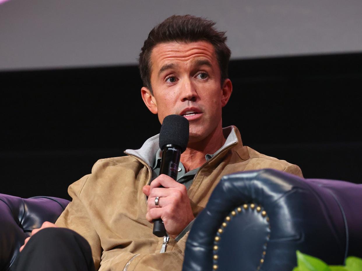 Rob McElhenney talks into a microphone while sitting on a large blue chair. He is wearing a tan quarter zip hoodie.