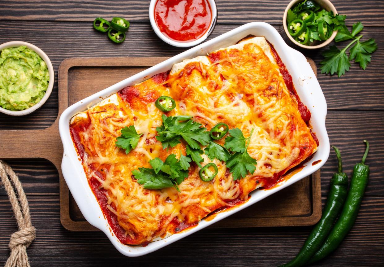 Traditional Mexican dish enchiladas with meat, chili red sauce and cheese in white casserole dish over rustic wooden background, served with guacamole and tomato dips. Close up, top view