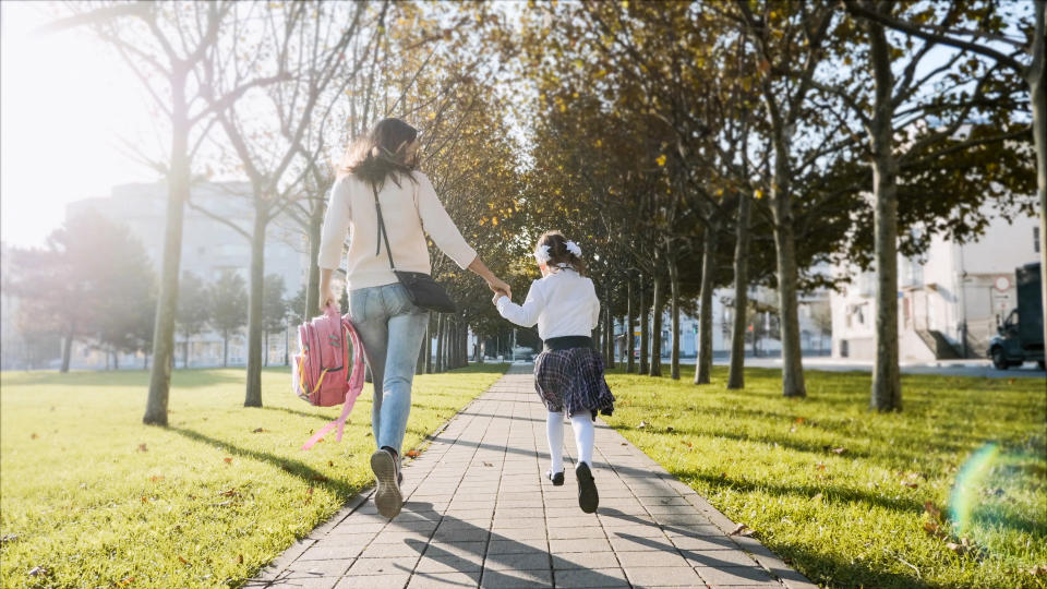 Young woman and little girl in school uniform are holding hands and running along the trees in the park at sunny autumn weather. Family late for school concept, back view.