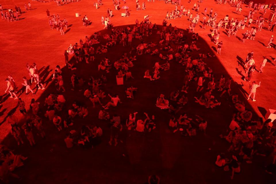 People seek some shade outdoors as others walk by in the sun in a red-tinted photo.