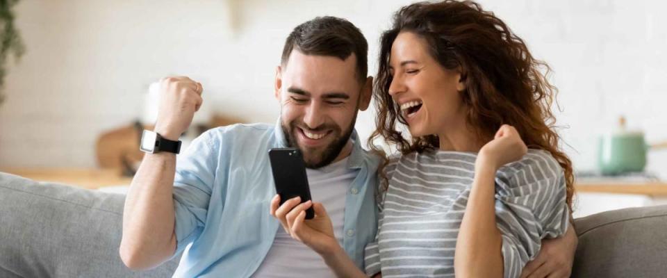 Excited overjoyed couple resting on couch holding smart phone, celebrating a low mortgage rate