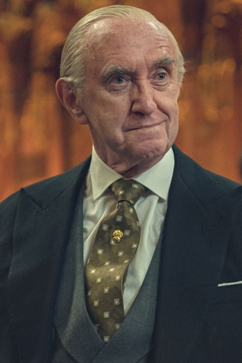 An older Jonathan Pryce in a suit and patterned tie stands in an ornate room with lamps and sculptures in the background