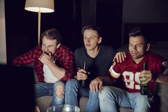 Three men, including one in a football jersey, sit on a sofa in front of a TV while drinking beer.