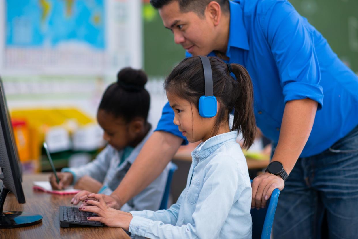 An elementary-school student wears headphones while working on a computer in class with the help of a teacher.