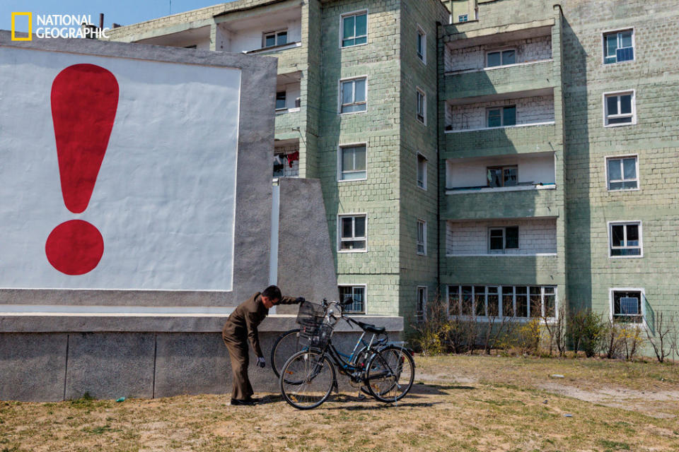 A man tends to his bicycle outside a housing complex in Kaesong, not far from the border with South Korea. An exclamation point at the end of an emphatic propaganda slogan punctuates the scene. (David Guttenfelder/National Geographic)