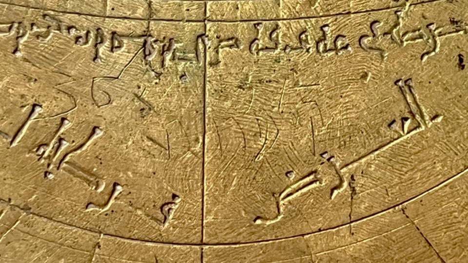 Κοντινό πλάνο ενός χάλκινου αστρονομικού οργάνου με αραβικά και εβραϊκά σημάδια και δυτικούς αριθμούς.