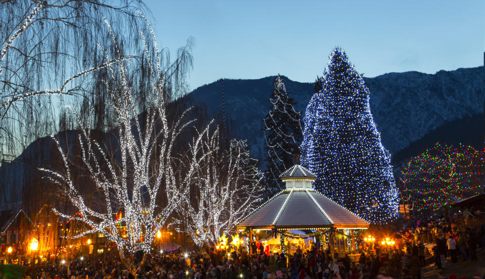 Festive holiday scene in a town square with a gazebo, large illuminated tree, and numerous people enjoying the atmosphere and decorations. Snow-capped mountains in the background