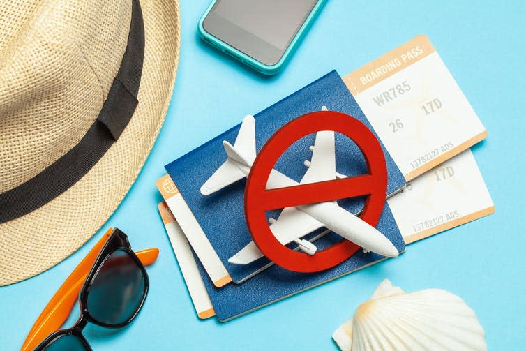 Hat, sunglasses, mobile phone, shell, two passports with boarding passes and a plane figurine with a no symbol over it