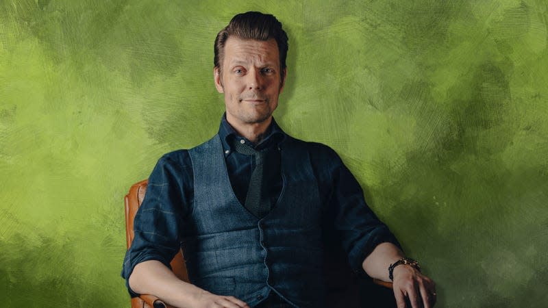 Sam Lake sits, snappily dressed in a vest and tie, against a green, splotchy background.