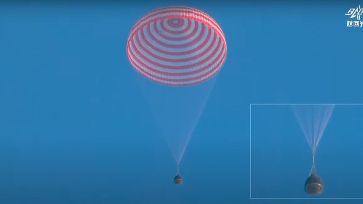  A brownish space capsule with a red-and-white-striped parachute descends through a blue sky. 