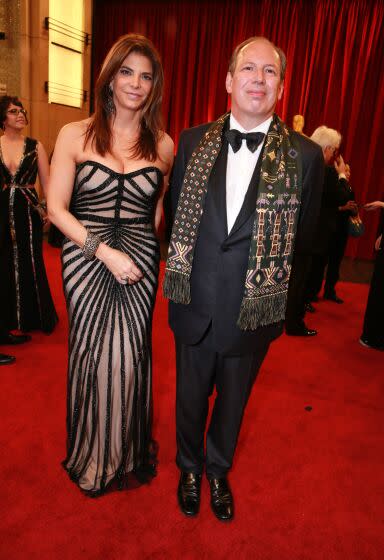 Dina De Luca is wearing a black, sheer, strapless dress alongside Hans Zimmer who is dressed in a black tuxedo and scarf