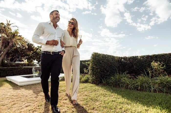 Well-dressed couple walking through greenery drinking champagne.