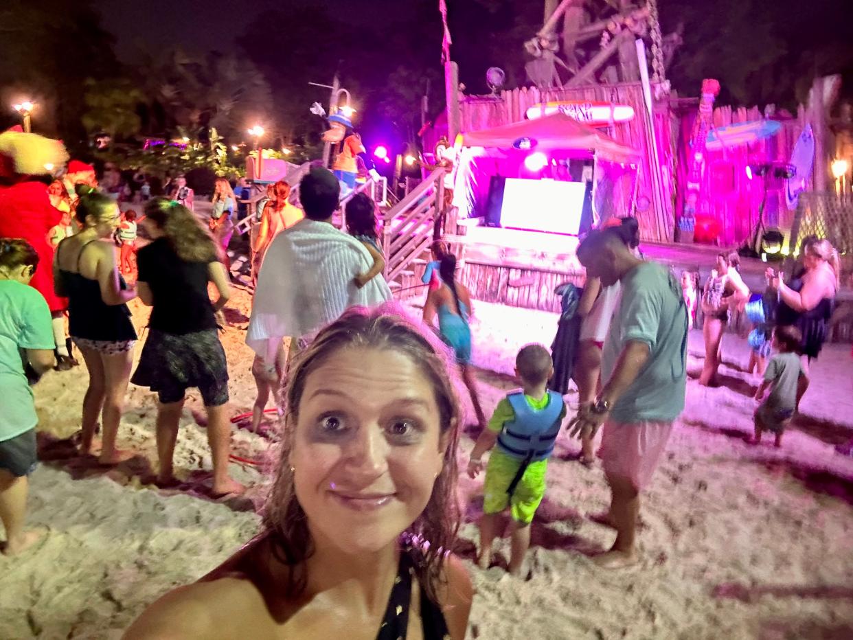 The writer smiling in front of groups of people dancing in front of lights in sandy area