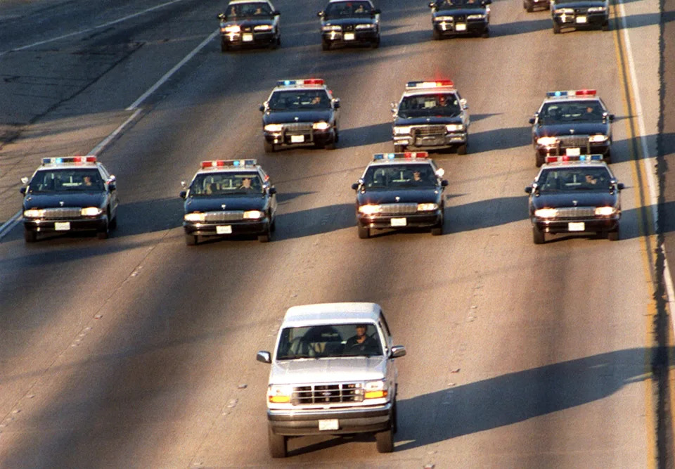 About a dozen police cars drive behind a white Ford Bronco across four highway lanes.