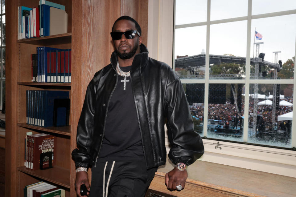 Sean "Diddy" Combs leans against a library window in a black leather jacket, dark sunglasses, and jewelry, with a crowd and stage visible outside