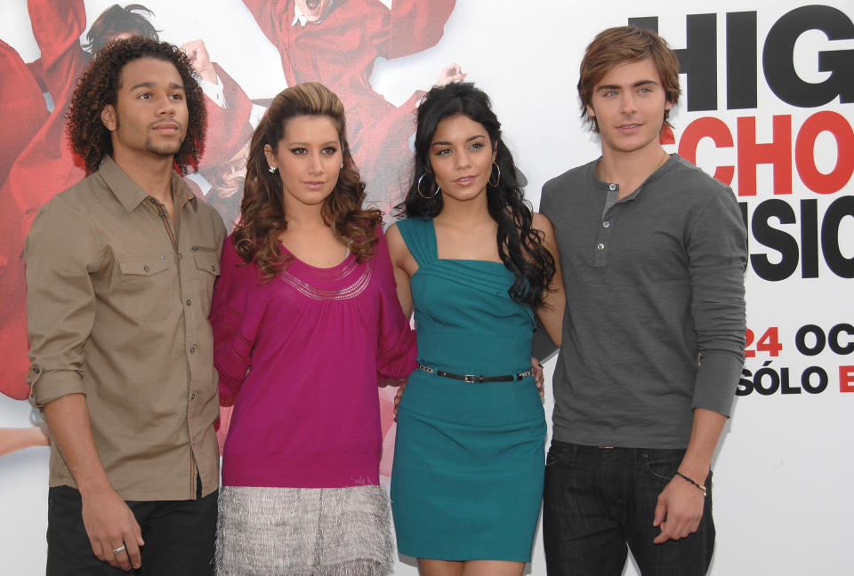 Corbin Bleu, Ashley Tisdale, Vanessa Hudgens, and Zac Efron stand together in casual outfits in front of a "High School Musical" poster