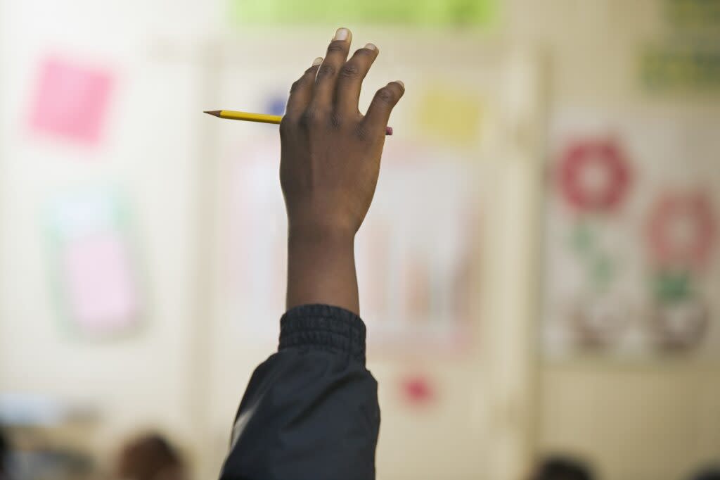 Student of color raising his hand showing only the hand holding a pencil in a classroom background