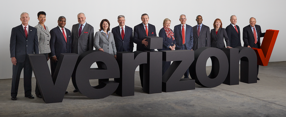 13 people in suits behind a Verizon sign.