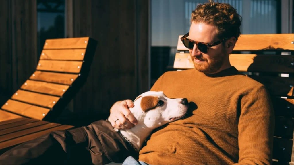 Man in sunglasses, pet sitting, dog on wooden bench – cheap travel hack.