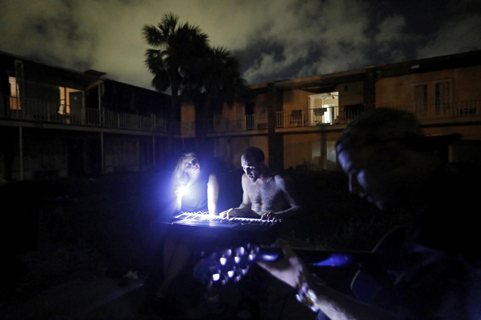 FILE - In this Oct. 16, 2018 file photo, Kevin Parker, center right, sits with his wife Lilith, while playing a song he wrote titled "My Life's Been Hell," on the keyboard while joined by neighbor Chris Thomas outside the damaged American Quality Lodge where they continue to live in the aftermath of Hurricane Michael, in Panama City, Fla. The tropical weather that turned into monster Hurricane Michael began as a relatively humble storm before rapidly blossoming into the most powerful cyclone ever to hit the Florida Panhandle, causing wrenching scenes of widespread destruction. (AP Photo/David Goldman, File)