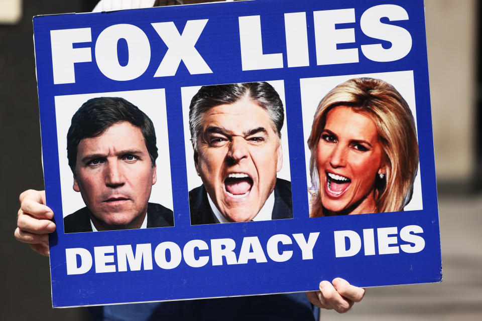 A Rise and Resist advocate holds a poster showing Carlson, Hannity and Ingraham, the last two of whom are shouting, marked: Fox Lies, Democracy Dies.