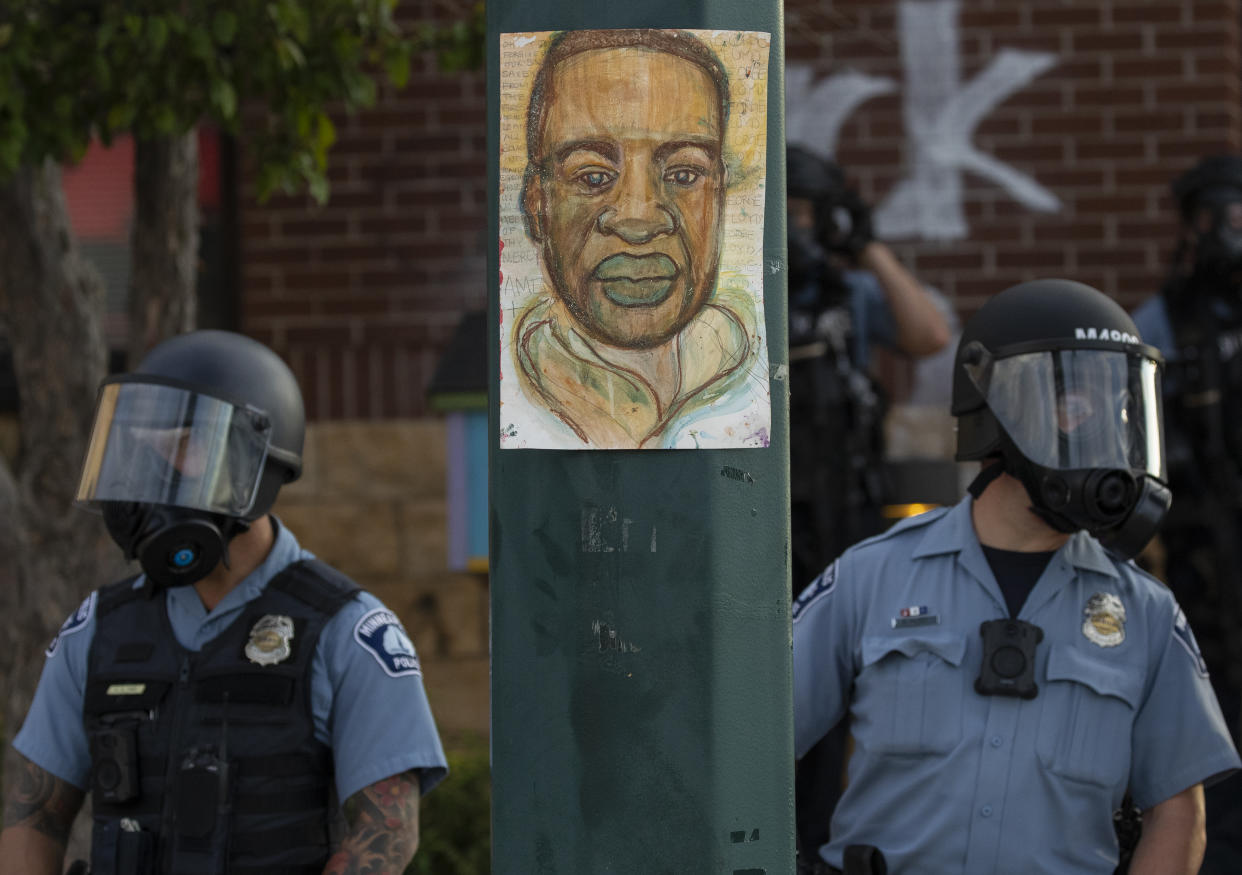 Two Minneapolis police officers wearing helmets with face shields stand alert next to a utility pole with an image of George Floyd posted on it.