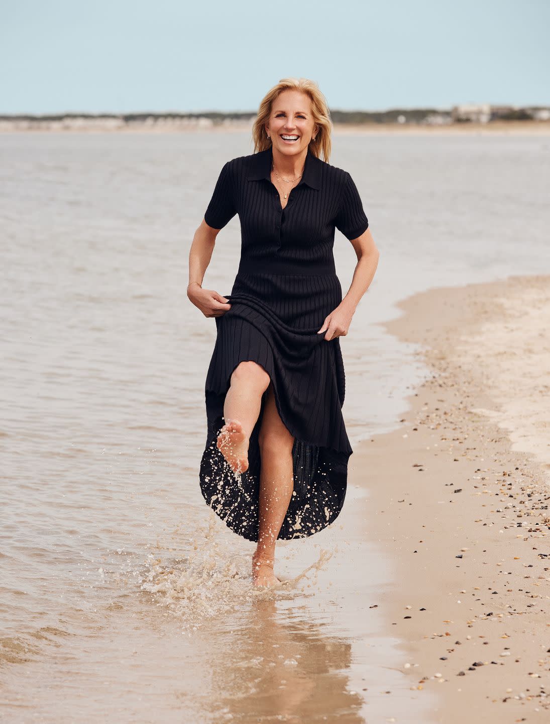 first lady jill biden in a black dress on the beach, kicking up water from the tide, smiling at the camera