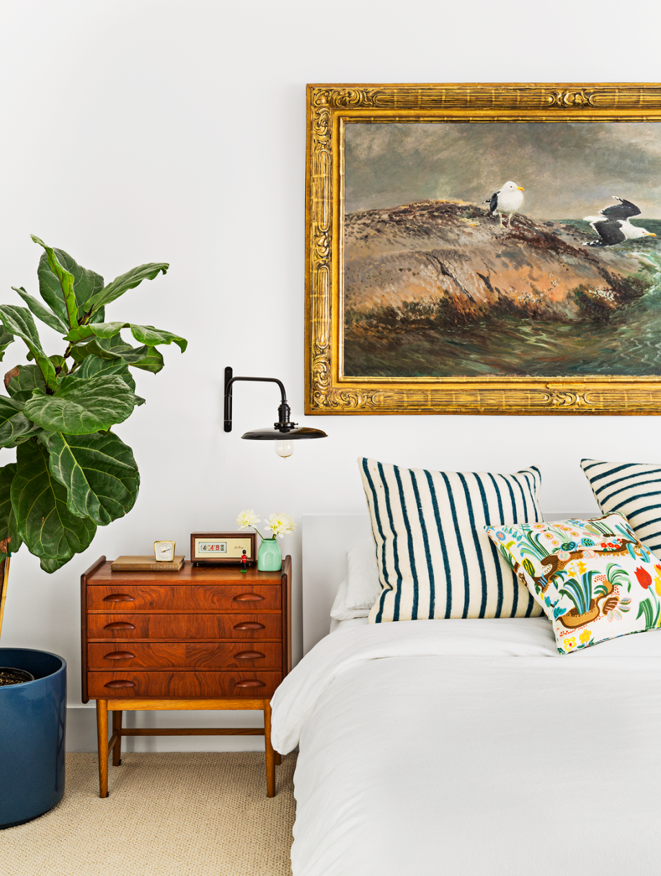 Master bedroom with a bright white bed with striped decorative pillows, a green potted plant, a midcentury wooden nightstand with a vintage clock, and a large framed mural depicting Birds on the Sea