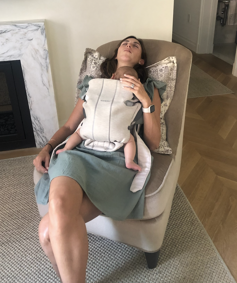 Hannah sleeping sitting in a chair with her legs crossed and Ellie in a carrier on her chest