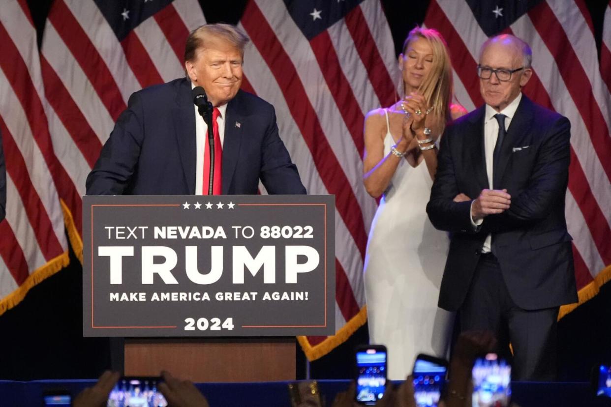 A man and woman stand by as Donald Trump speaks at a lectern with a campaign sign, in front of several draped American flags