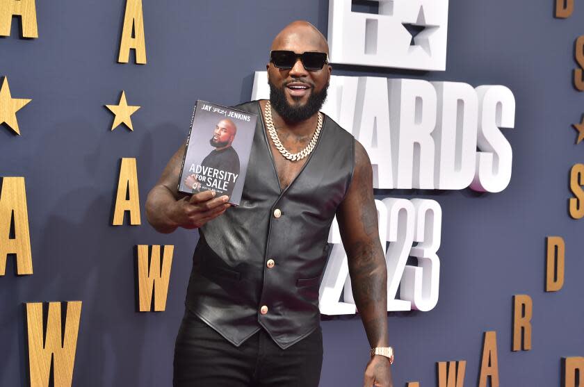 Jeezy is posing with his memoir, "Adversity for Sale," and is smiling while wearing sunglasses and a black vest