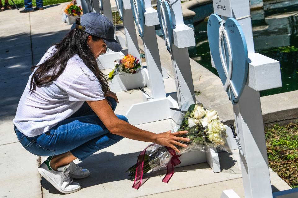 Meghan Markle in a black cap, white t shirt and jeans, crouches down with a bouquet of white roses at a Texas school shooting memorial. 