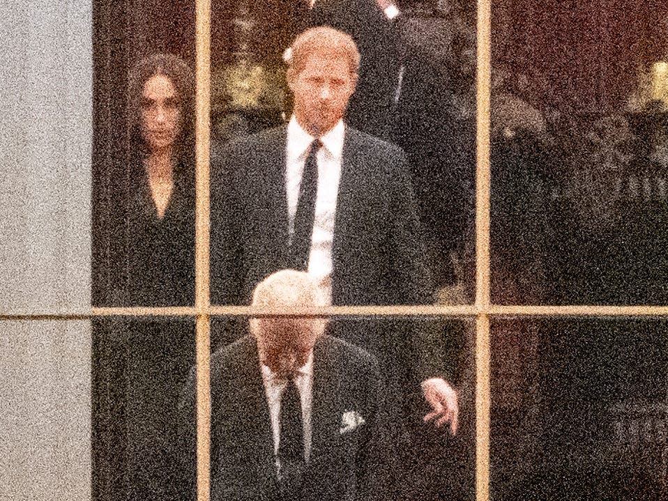 A close-up photo shows Meghan Markle, Prince Harry, and King Charles through a window of Buckingham Palace.