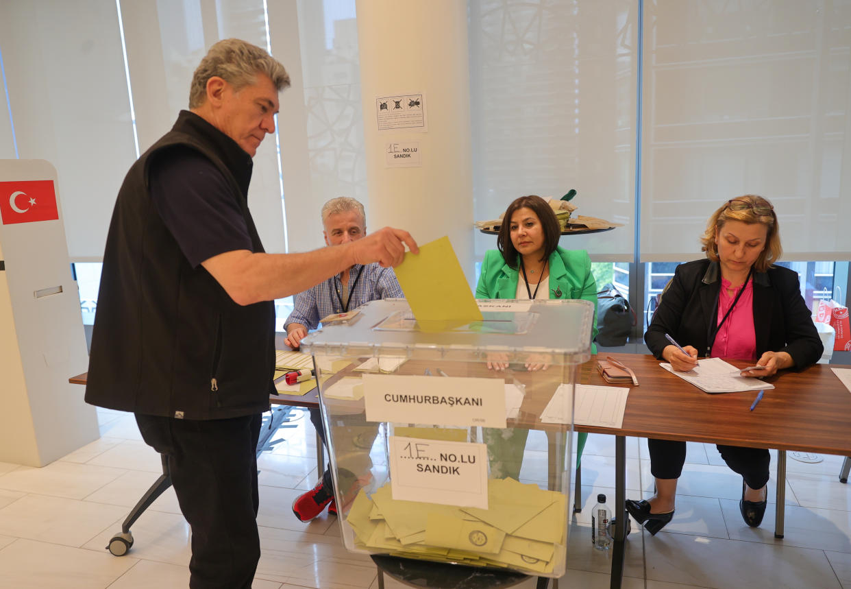 A middle-aged man puts a yellow ballot through a slot into a clear plastic bucket, with three election staff sitting at tables behind him.