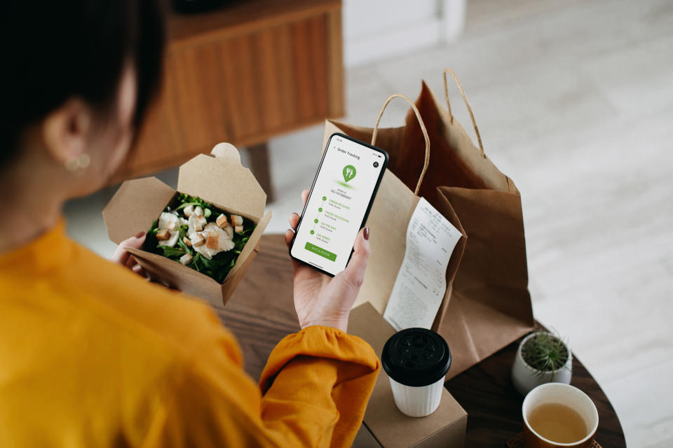 Over the shoulder view of young Asian woman ordering home delivery takeaway food with mobile app on smartphone, tracking status and confirming the food order. Enjoying a box of fresh grilled chicken vegetable salad. Technology makes life so much easier