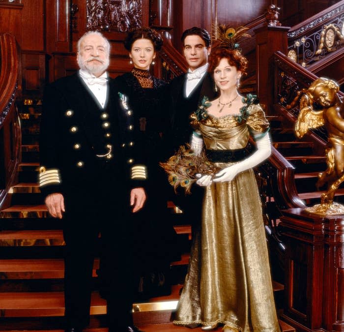 Four characters from the movie "Titanic" are posing in front of a grand staircase; they are dressed in elegant period costumes