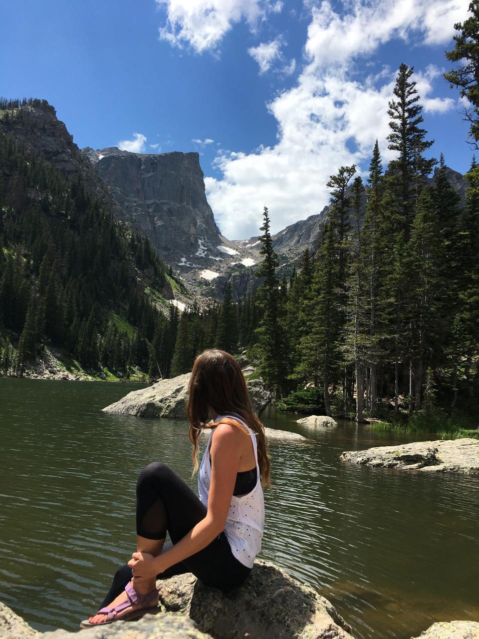 Emily, wearing purple sandals, black leggings, and a black and white tank top, sitting on a rock and staring out at the mountains and trees on a lake. 