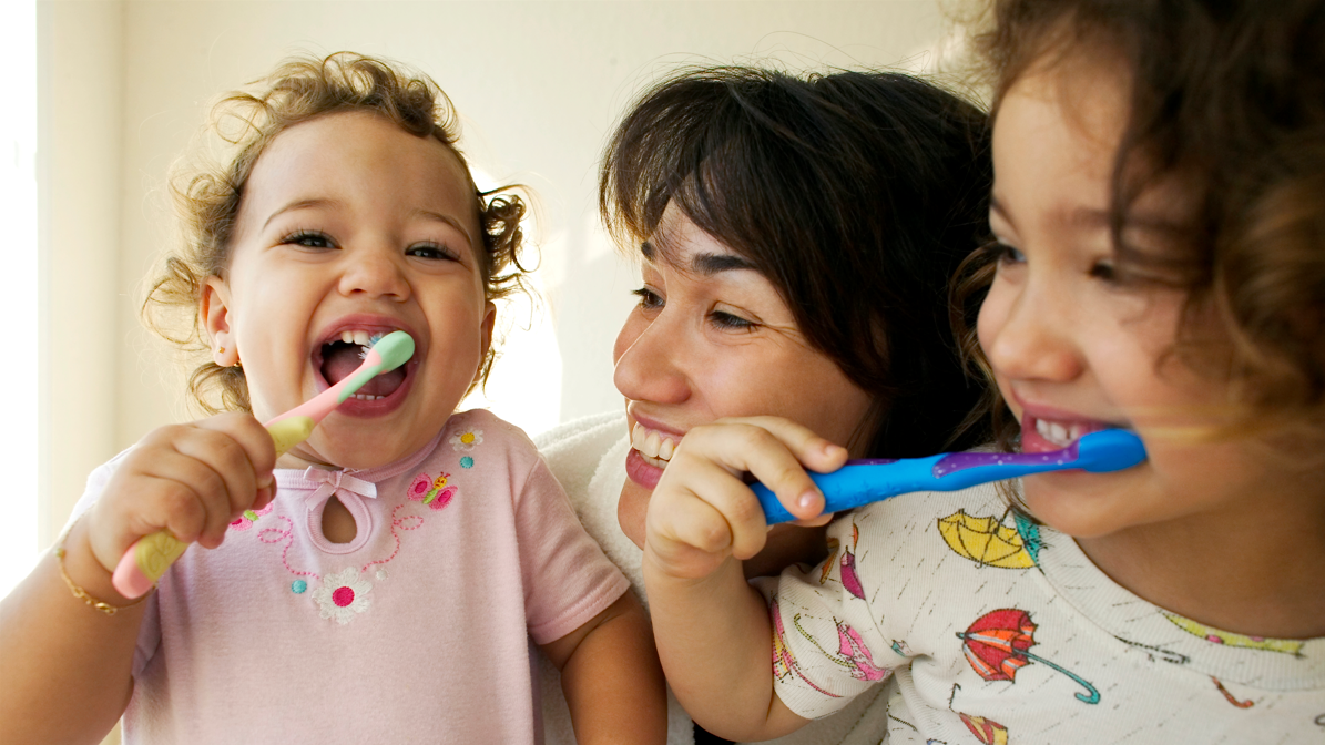 A woman smiles at two grinning children brushing their teeth