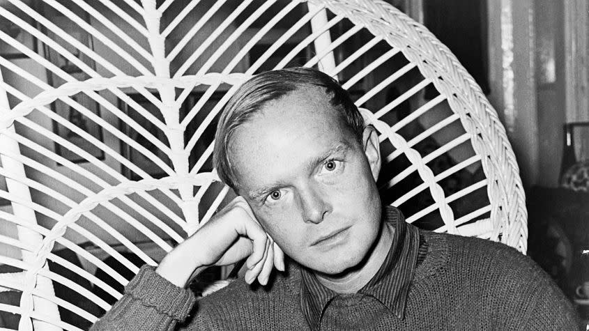 truman capote sits in a highbacked wicked chair and looks at the camera as he rests his head on one hand, he wears a sweater over a collared shirt and slacks