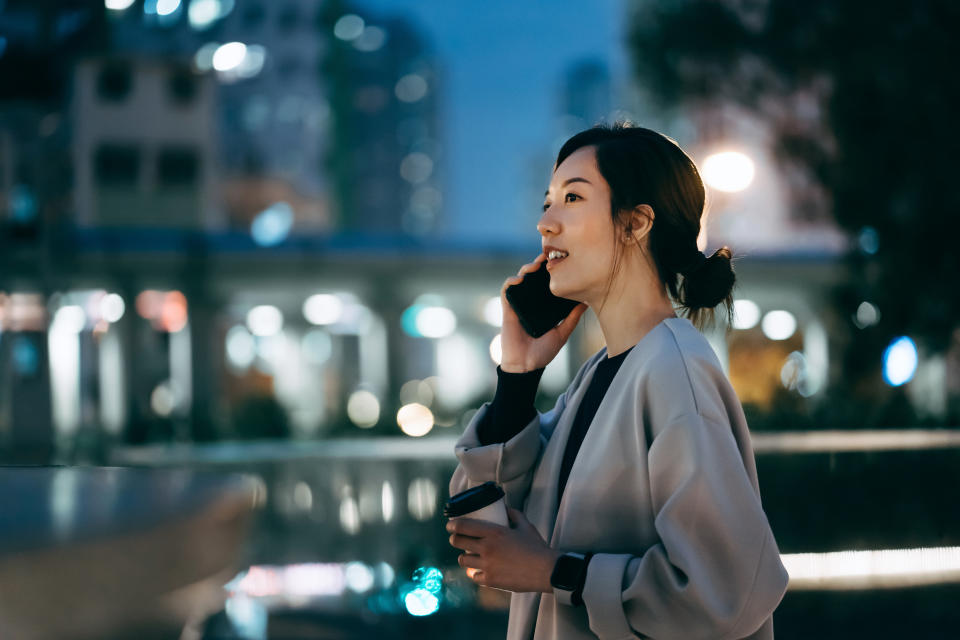 Confidence young Asian businesswoman having a cup of coffee, talking on smartphone on the go while walking in downtown city street, against illuminated urban city scene after work in the evening