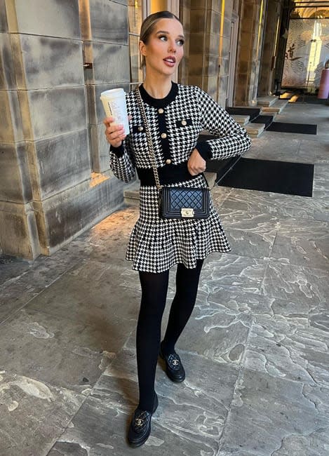 a full length photo of helen flanagan standing with one hand on her hip and the other clutching a paper coffee cup wearing a tightly fitted black and white houndstooth dress which has gold buttons running from the neckline to waistline and she wears chunky flat black shoes with a black crossbody handbag