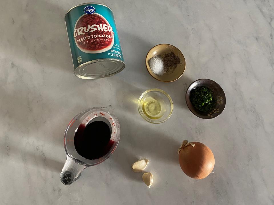 Can of crushed tomatoes, onion, cloves of garlic, a liquid measuring cup filled with red wine, oil, and spices on a table