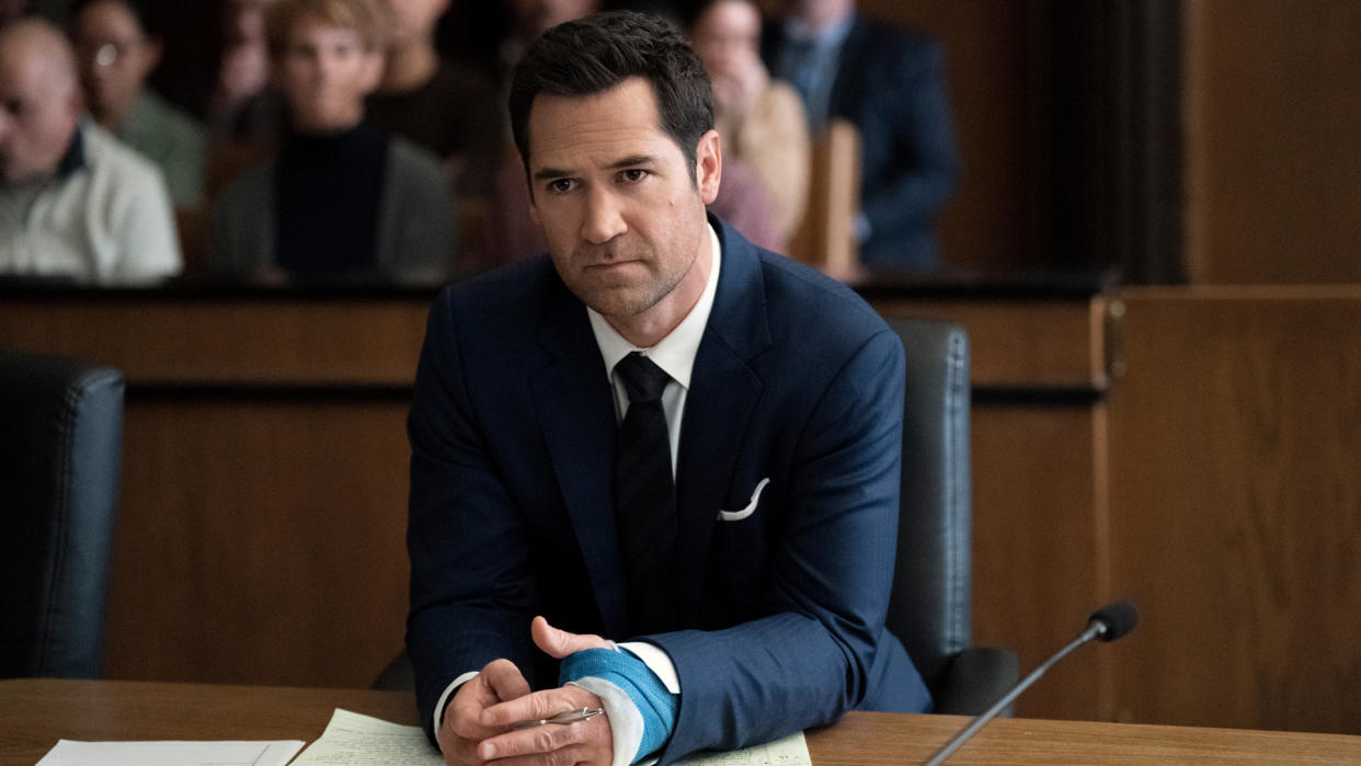  Mickey Haller sits at a desk in a court room with a bandaged hand in The Lincoln Lawyer season 2, which precedes The Lincoln Lawyer season 3 