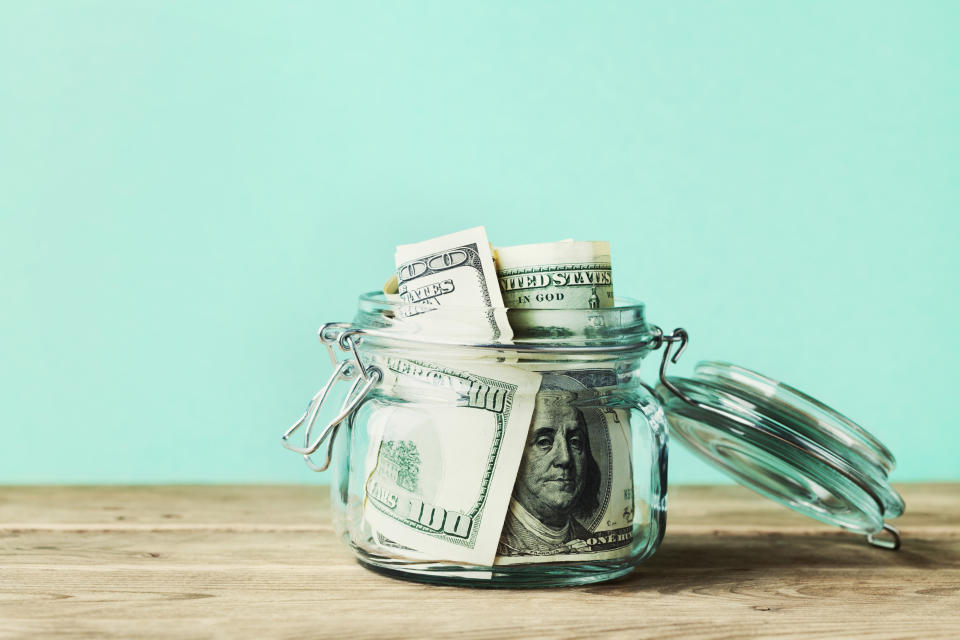 A clear glass jar filled with neatly folded U.S. dollar bills, placed on a wooden surface against a plain background